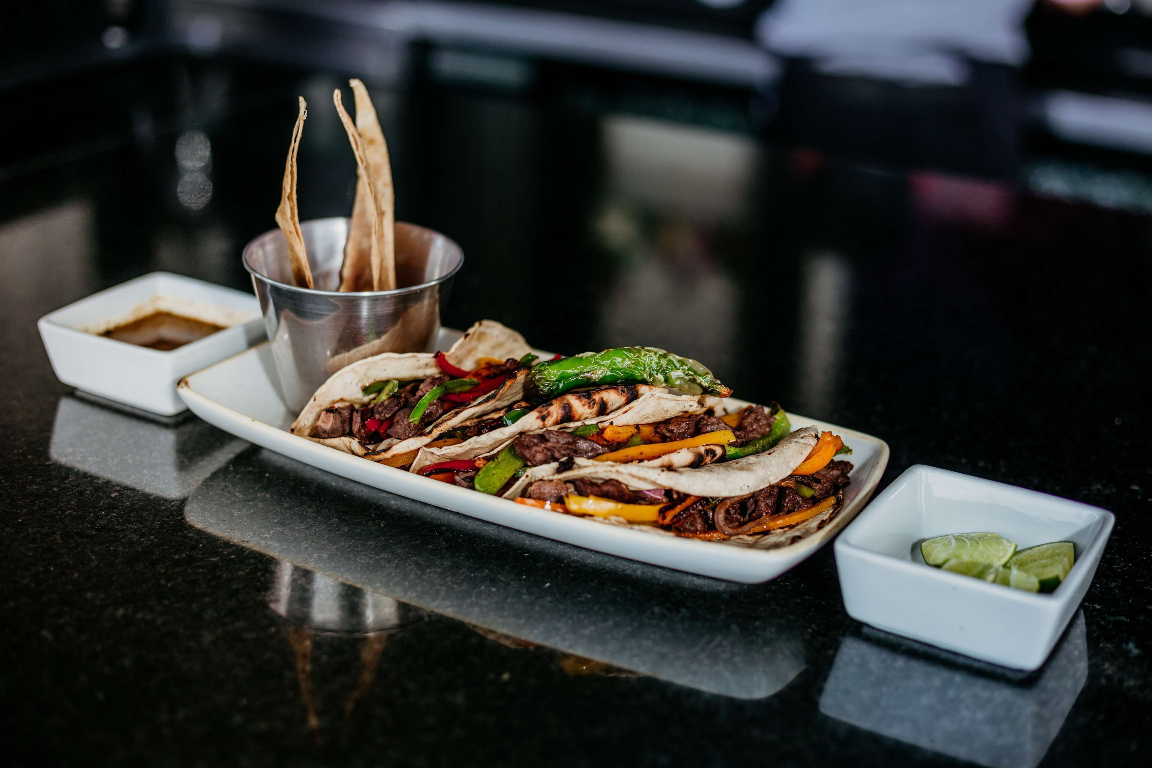 A plate of tacos with various toppings and two small bowls of sauces on a black countertop.