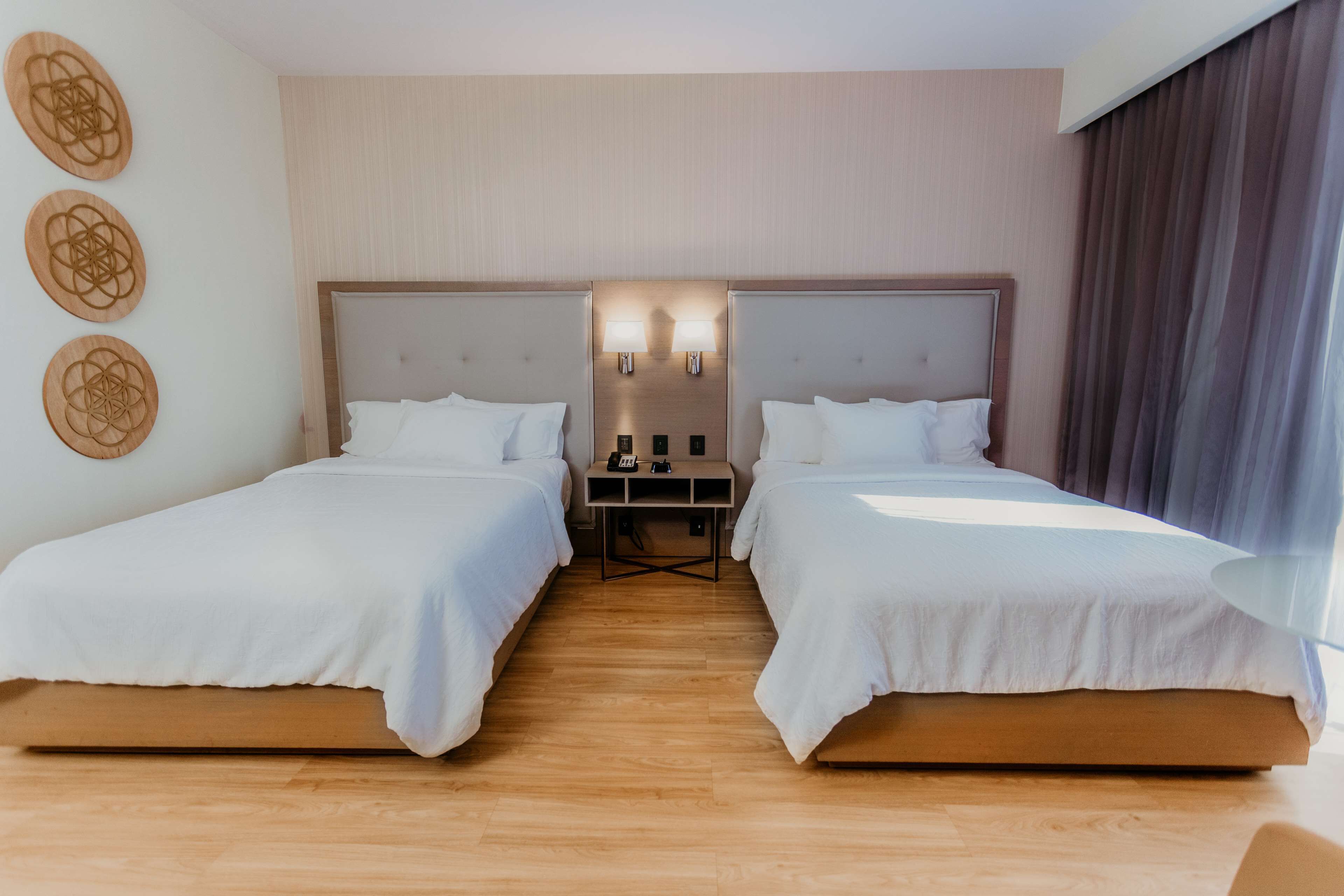 Une chambre d’hôtel avec deux lits simples, un plancher en bois, une table de chevet avec une lampe et un mur orné de trois motifs circulaires.