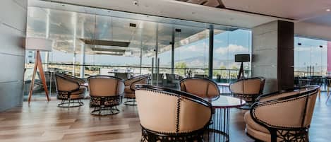 Un área de estar interior moderna con paredes de vidrio, techo de madera y vista a un puente.