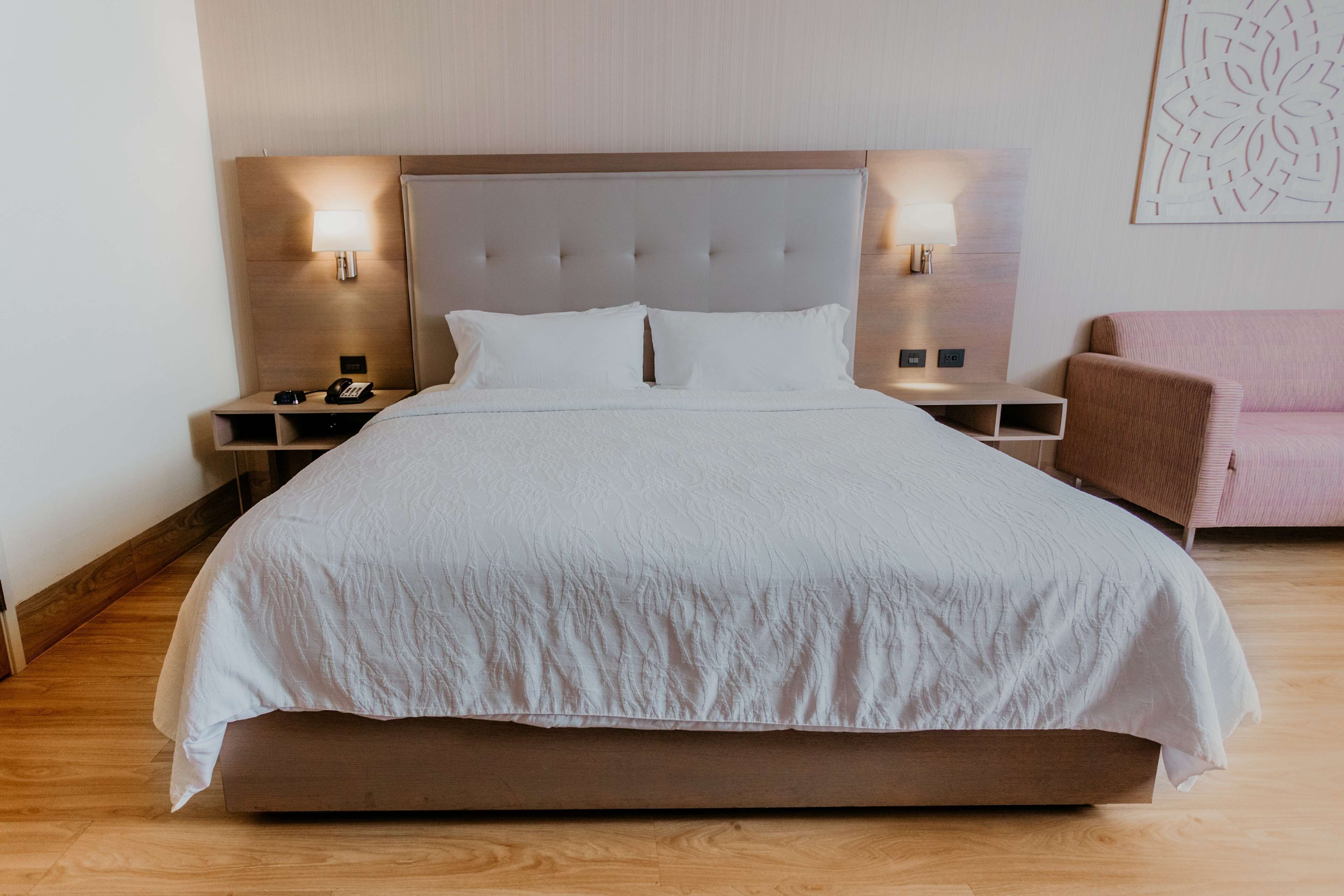 Una cama grande con cabecera capitonada, dos mesitas de noche con lámparas, y un sillón rosa.