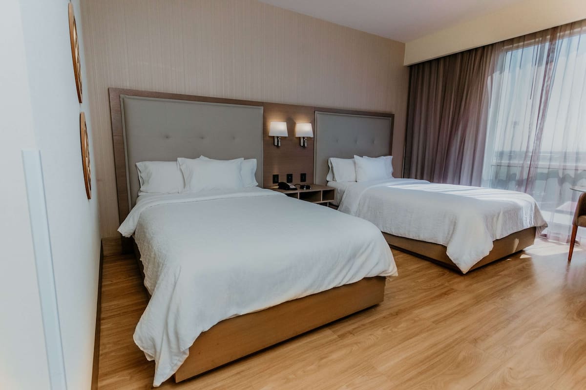 Habitación de hotel con dos camas, suelo de madera y un ventanal con cortinas.