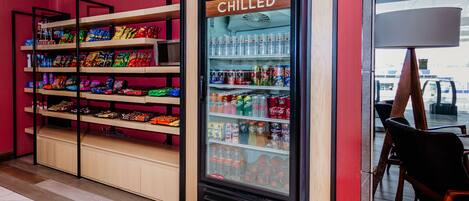 A chilled beverage display in a store with shelves of snacks and a sign reading "The Shop".