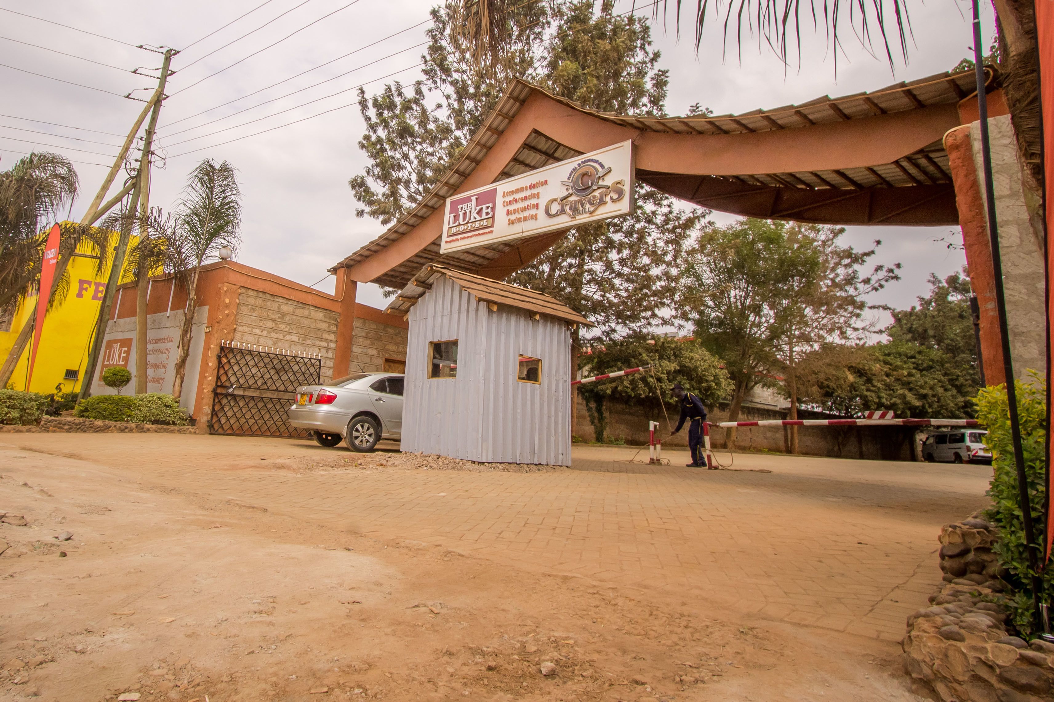 A gated entrance with a sign for 'The Luke' and a small building, a car parked outside, and a person standing near the gate.