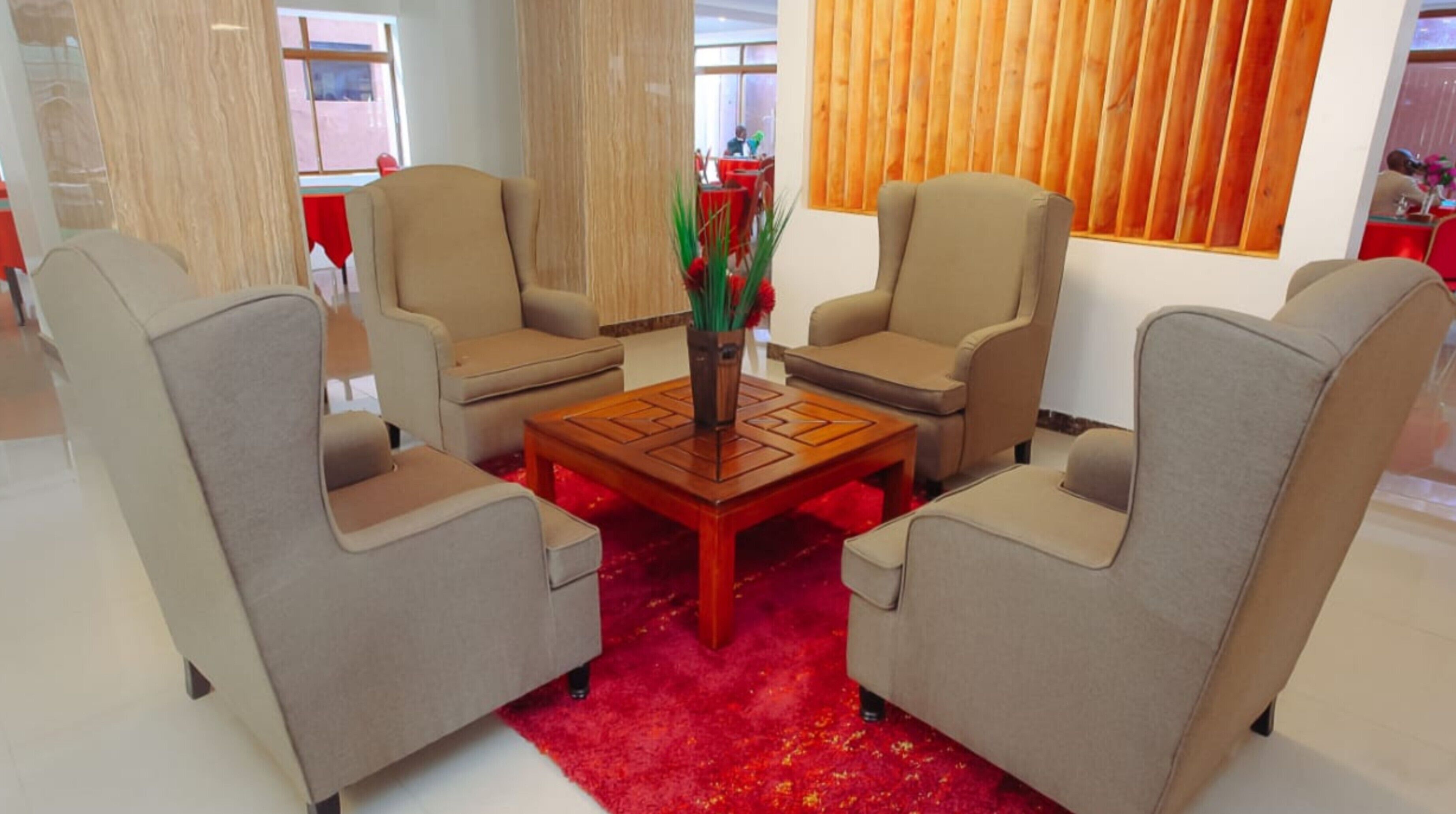 Un área de descanso con cuatro sillones beige y una mesa de centro de madera, que incluye una alfombra roja y un jarrón con plantas verdes.