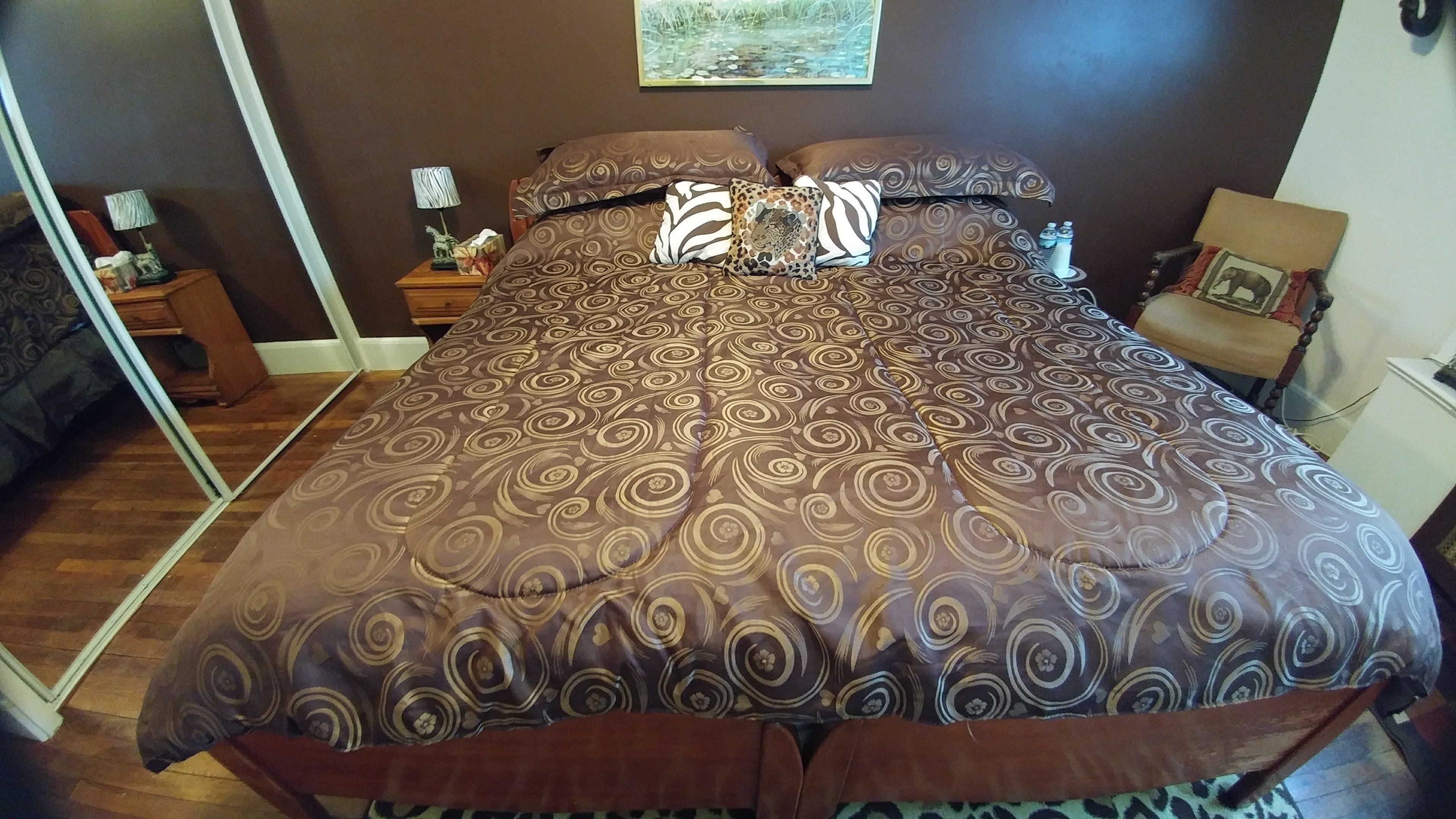 Un dormitorio con una cama grande, ropa de cama estampada, una mesita de noche con una lámpara y un cuadro en la pared.