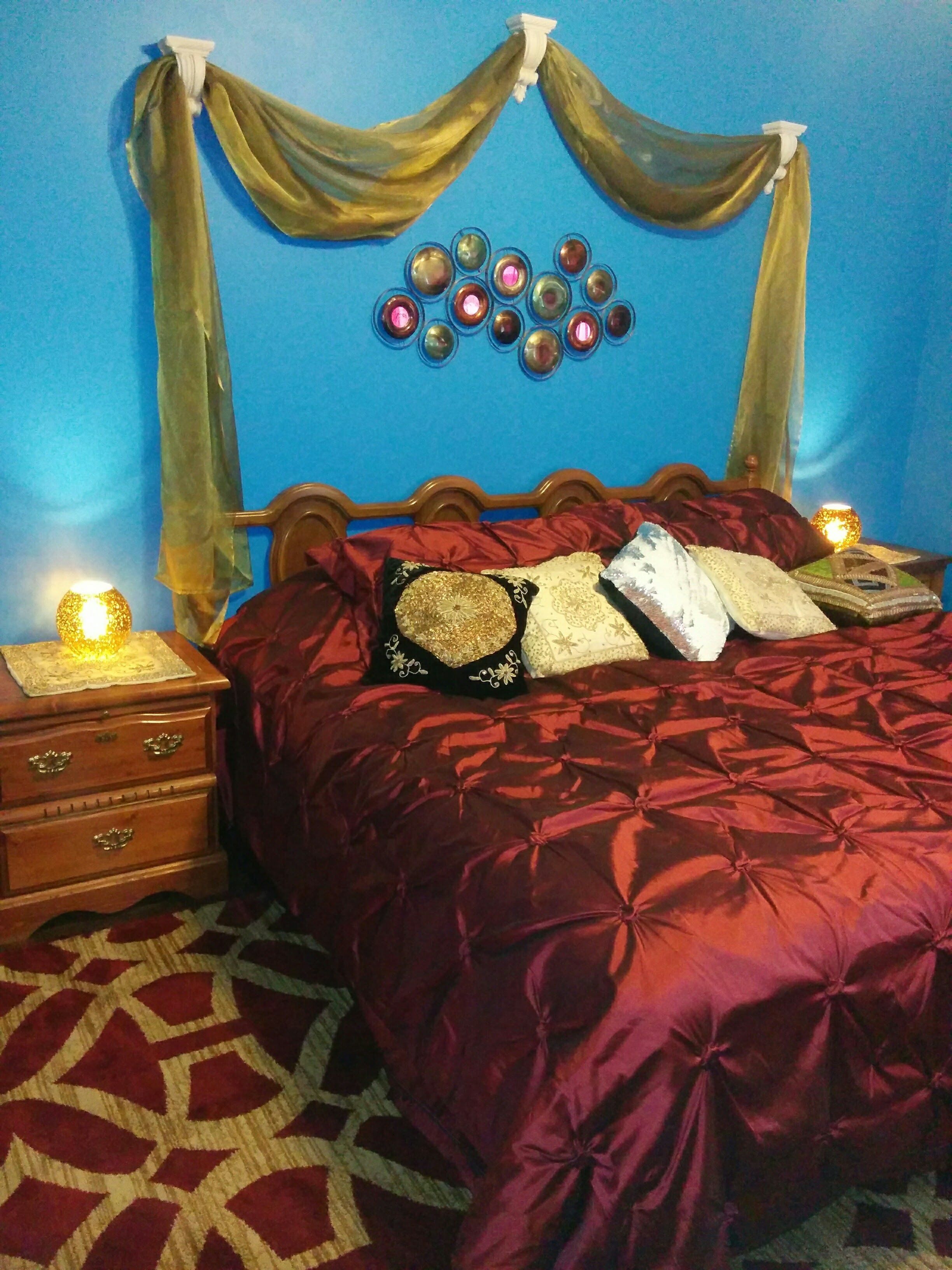 Un dormitorio con cama de madera, ropa de cama roja, una alfombra estampada, una mesita de noche con lámpara y una pared decorada.