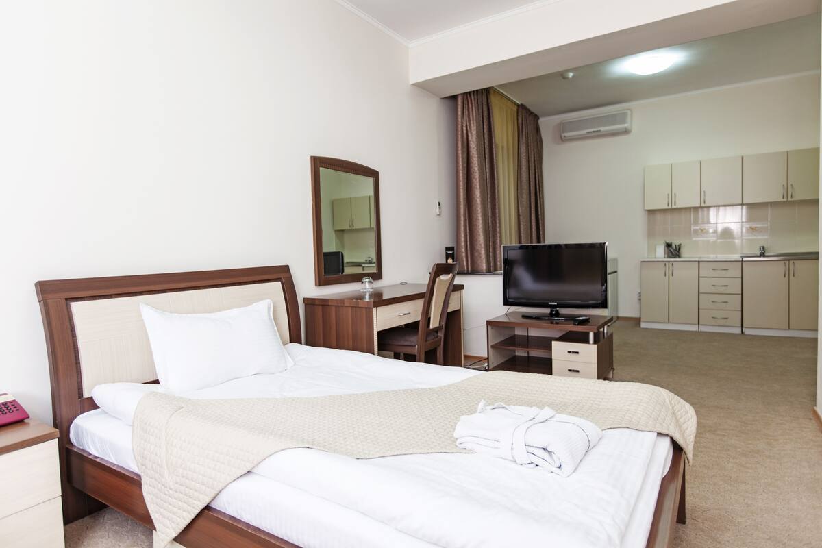 A hotel room with a bed, a desk with a television, a mirror, and a kitchenette area.