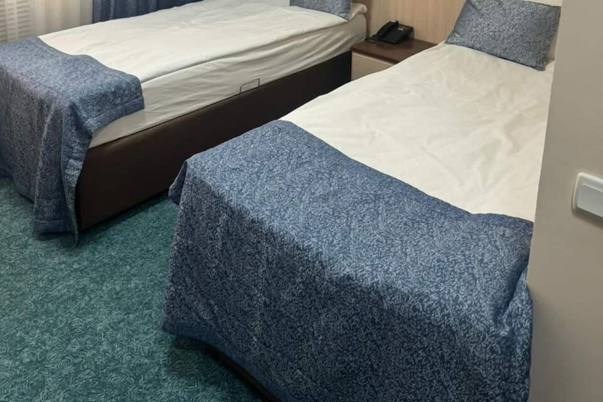 A hotel room with two beds, a window with curtains, an air conditioning unit, and a bedside table with a phone.