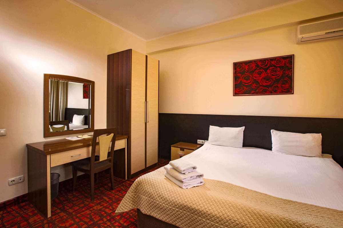 A hotel room with a bed, a desk, a chair, a mirror, a nightstand, and a wall-mounted artwork.