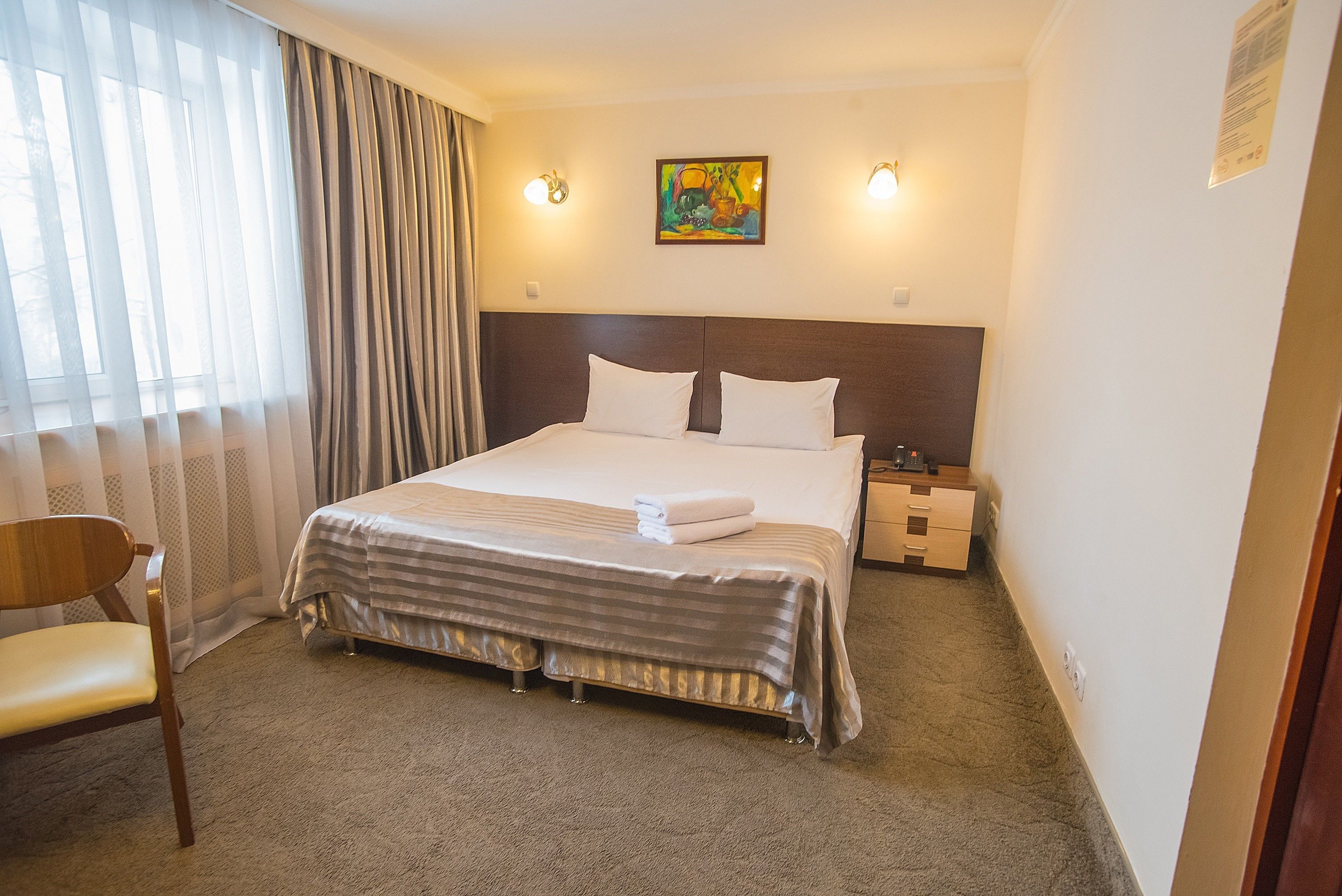 A hotel room with a large bed, a nightstand, a chair, and a painting on the wall.