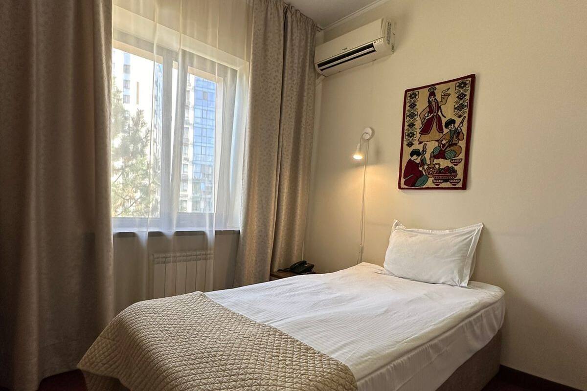A hotel room with a bed, a window with curtains, an air conditioning unit, a wall-mounted lamp, and a decorative piece.