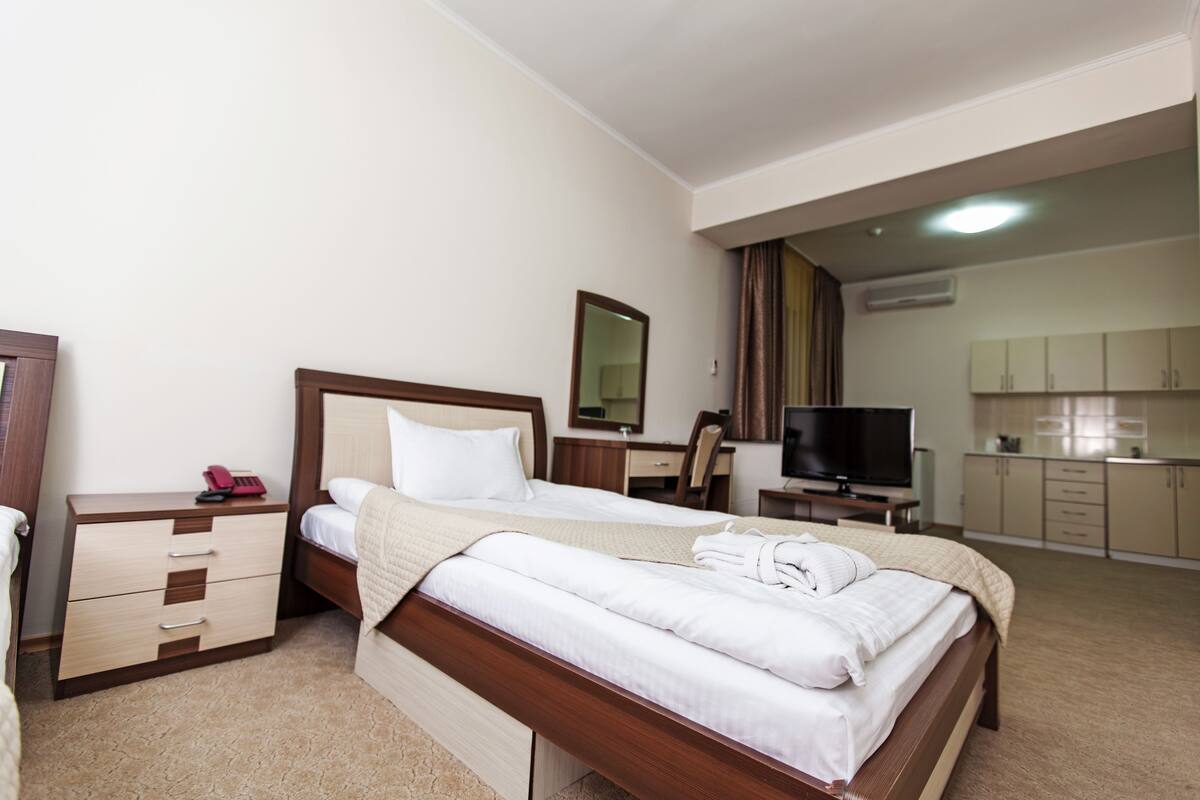 A hotel room with a bed, bedside table, TV, and a kitchenette area.