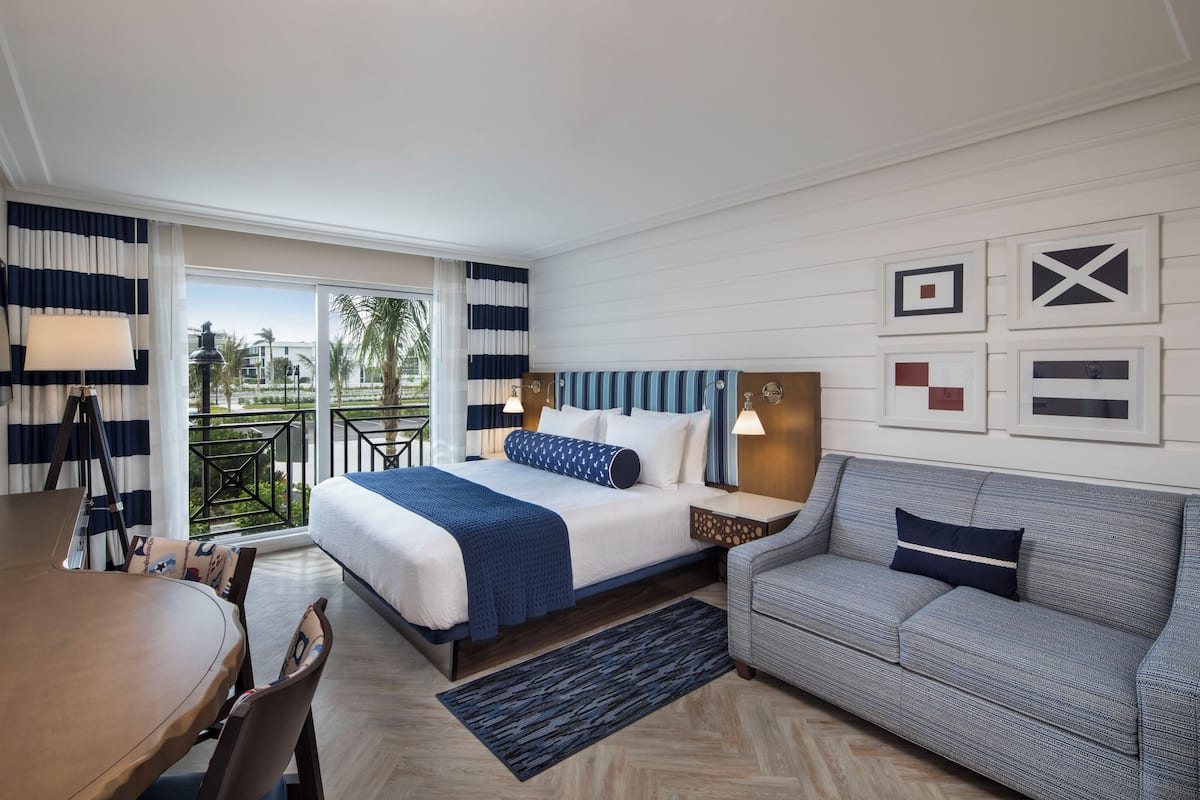 A hotel room with a bed, a sofa, a dining table, and a balcony with a view.
