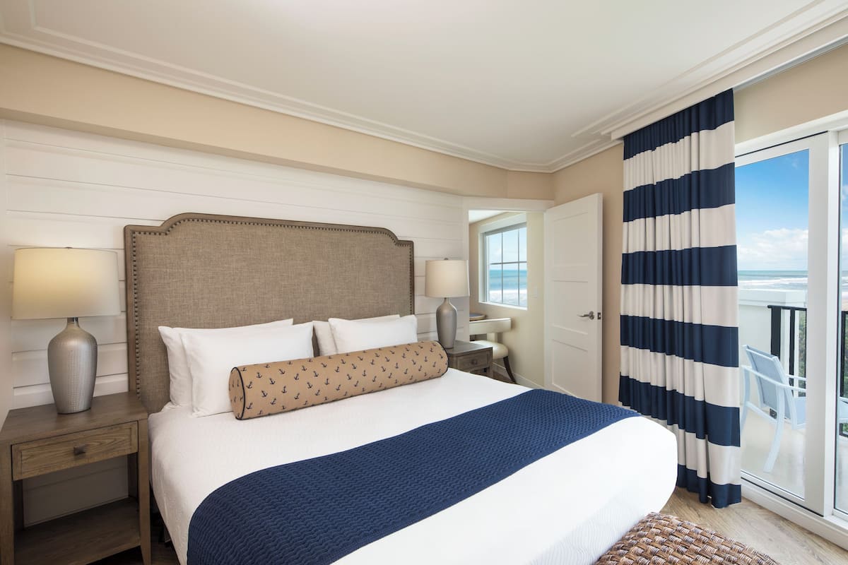 A hotel room with a large bed, a nightstand, a lamp, and a view of the ocean through a sliding glass door.