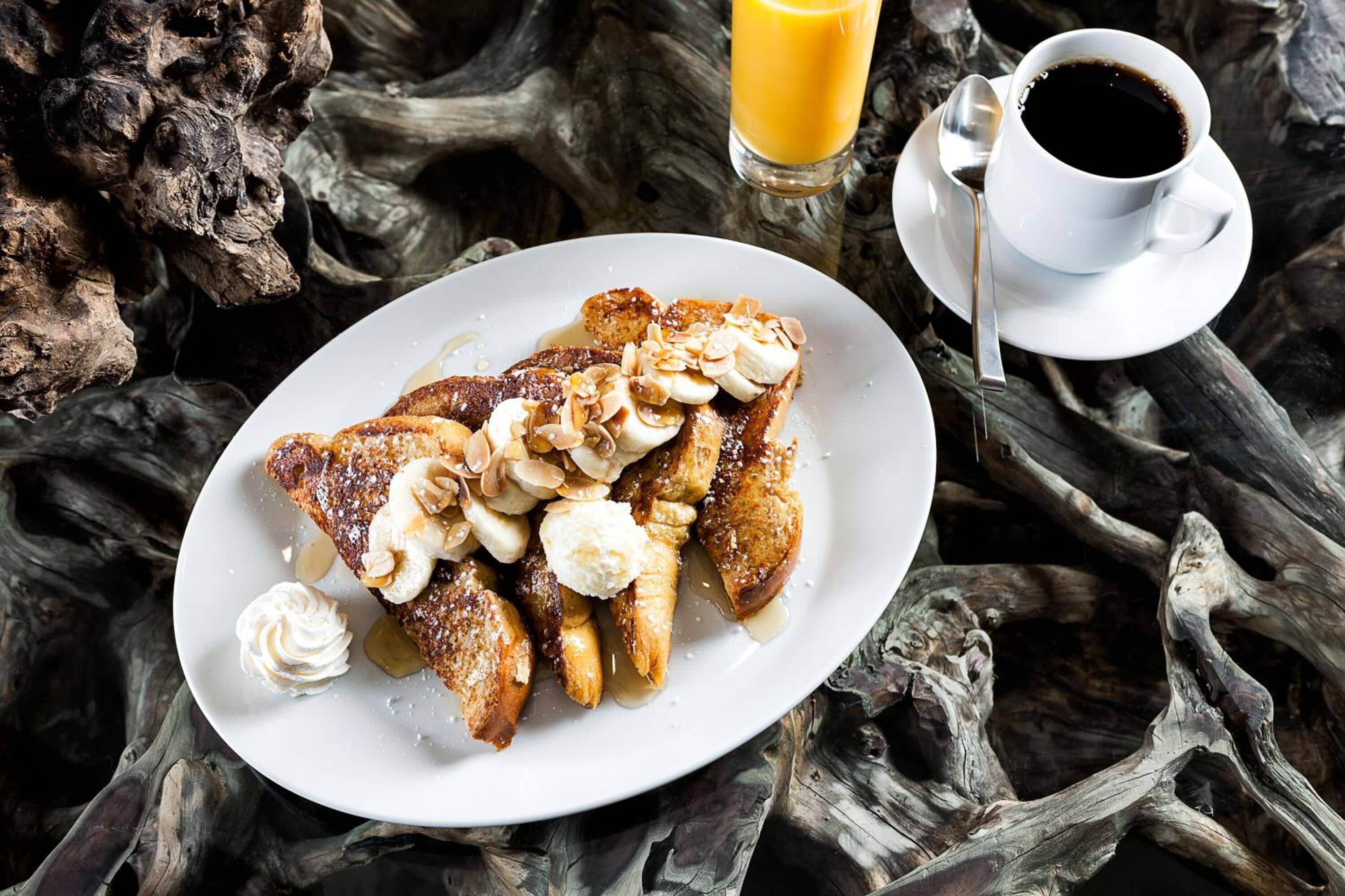 A plate of French toast with almonds and whipped cream, a glass of orange juice, and a cup of coffee on a rustic wooden table.