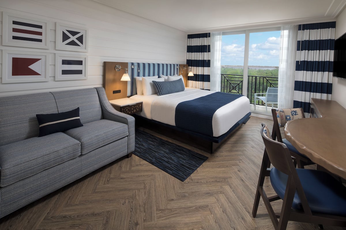 A hotel room with a bed, a sofa, a dining table, and a balcony with a view.