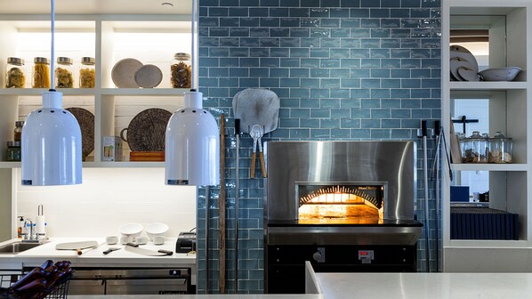 A modern kitchen with a wood-fired oven, hanging light fixtures, and shelves with jars and kitchenware.