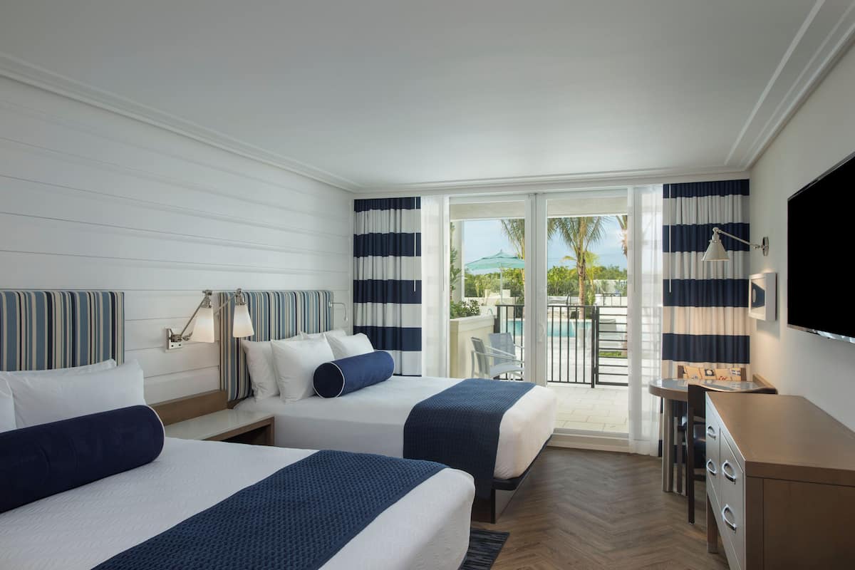 A hotel room with two beds, a desk, a TV, and a balcony with a pool.