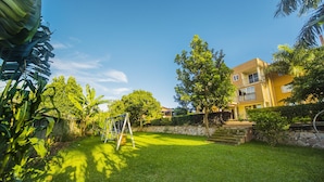 A well-maintained backyard with a swing set, a stone wall, and a multi-story building in the background. - Hotel Royal Nest (Entebbe)