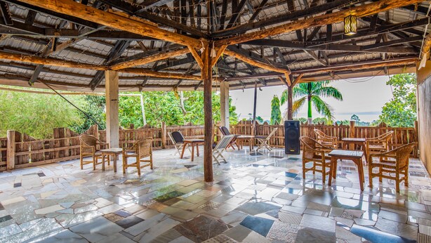 An outdoor covered patio with tiled flooring, wooden furniture, and a view of greenery and palm trees. - Hotel Royal Nest (Entebbe)