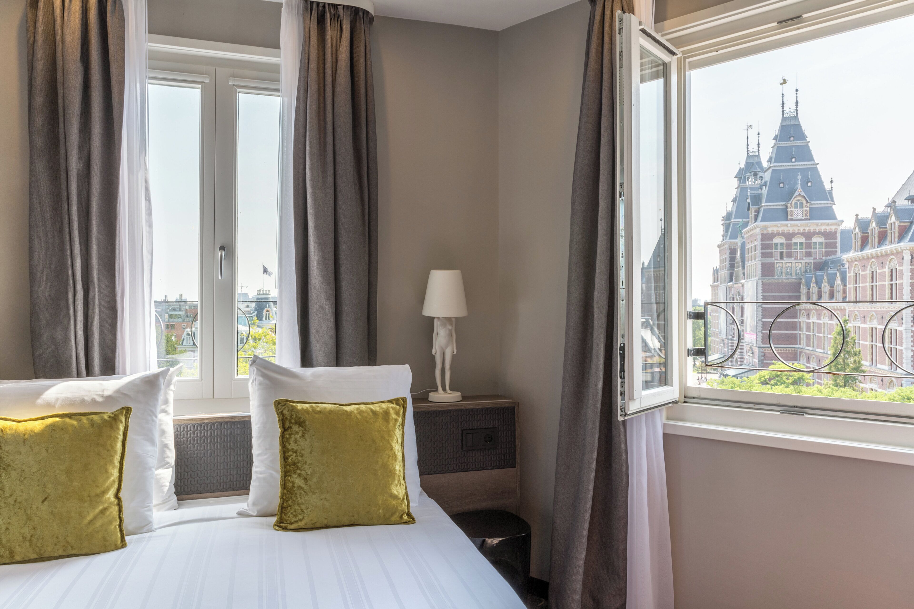 Habitación de hotel con cama, cortinas, mesita de noche, lámpara y ventana con vista a un edificio.