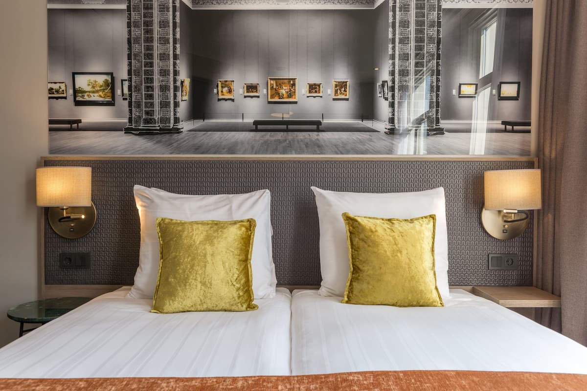 Hotellihuone, jossa on suuri sänky, kaksi kultaista koristetyynyä, yöpöytä lampun kanssa ja suuri ikkuna, josta avautuu näkymä taidegalleriaan.