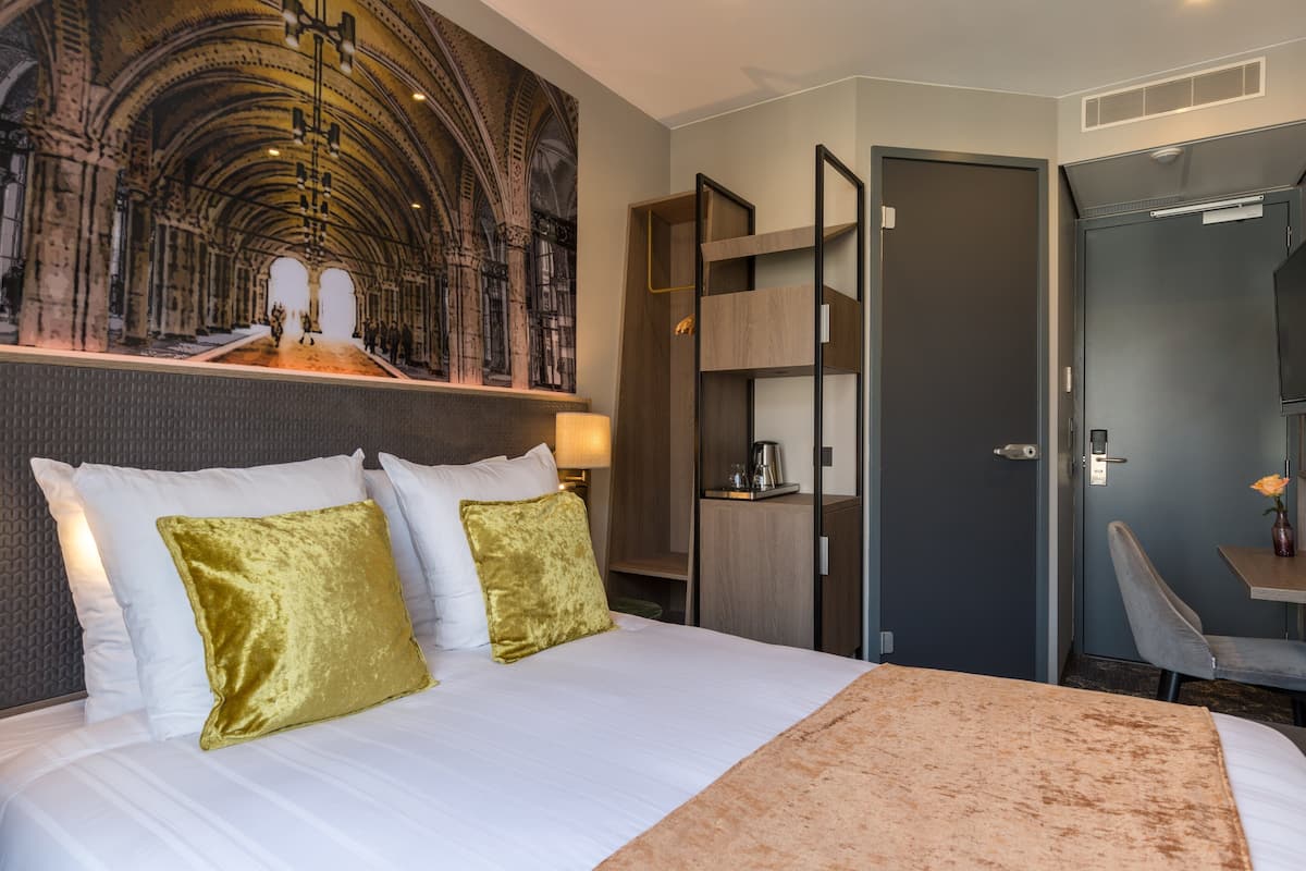 Hotellihuone, jossa on suuri sänky, kaksi kultaista tyynyä, puinen pääty, työpöytä tuolilla ja suuri seinämaalaus.