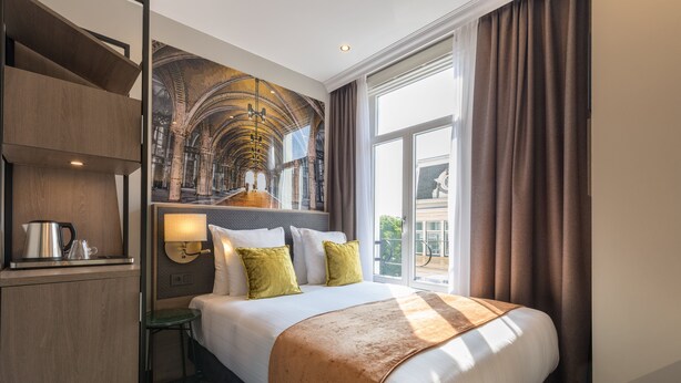 Habitación de hotel con una cama grande, un escritorio con hervidor, una mesita pequeña y una lámpara de pared.