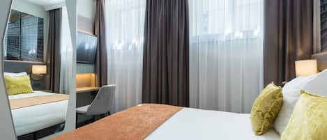 Habitación de hotel con cama, escritorio, silla, espejo grande y ventana con cortinas.