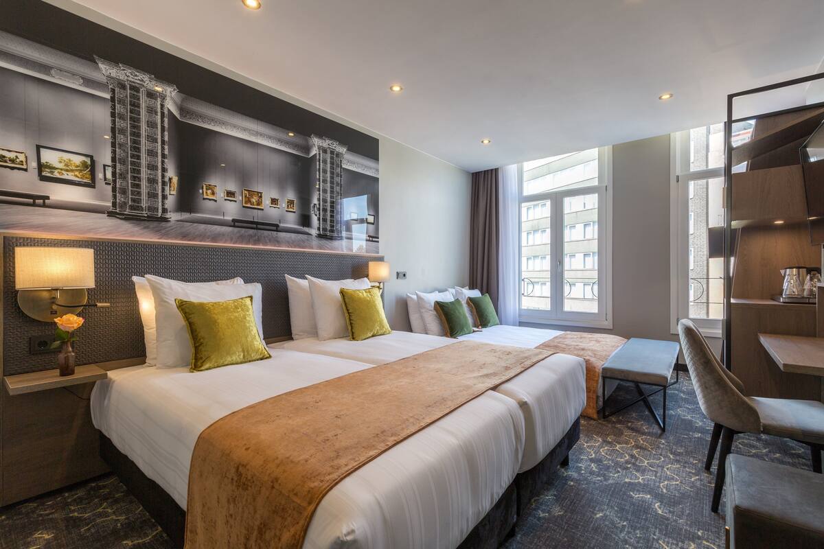 Une chambre d’hôtel avec deux lits, un bureau et une chaise. On y trouve des lampes fixées au mur et des œuvres d’art.