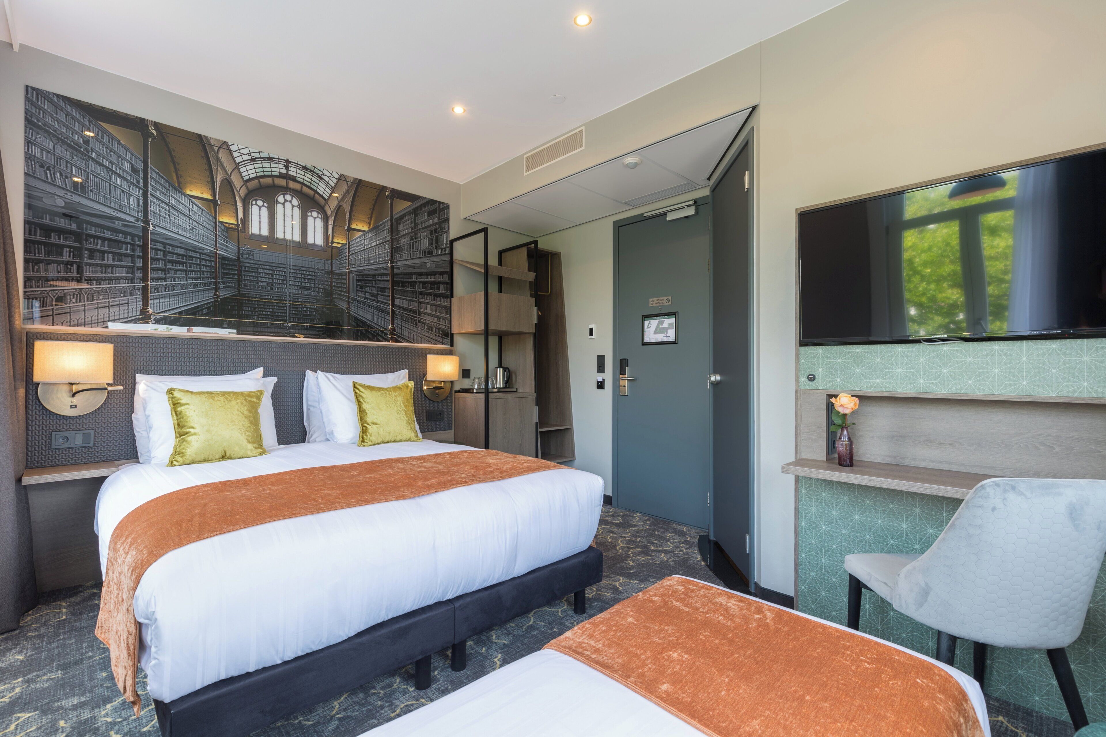 Una habitación de hotel con una cama grande, un escritorio, una silla, un televisor y un mural que representa una biblioteca.