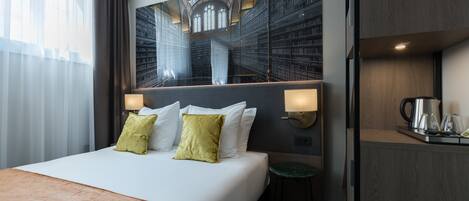 Habitación de hotel con una cama grande, un escritorio con tetera y vistas a una biblioteca a través de una pared de vidrio.