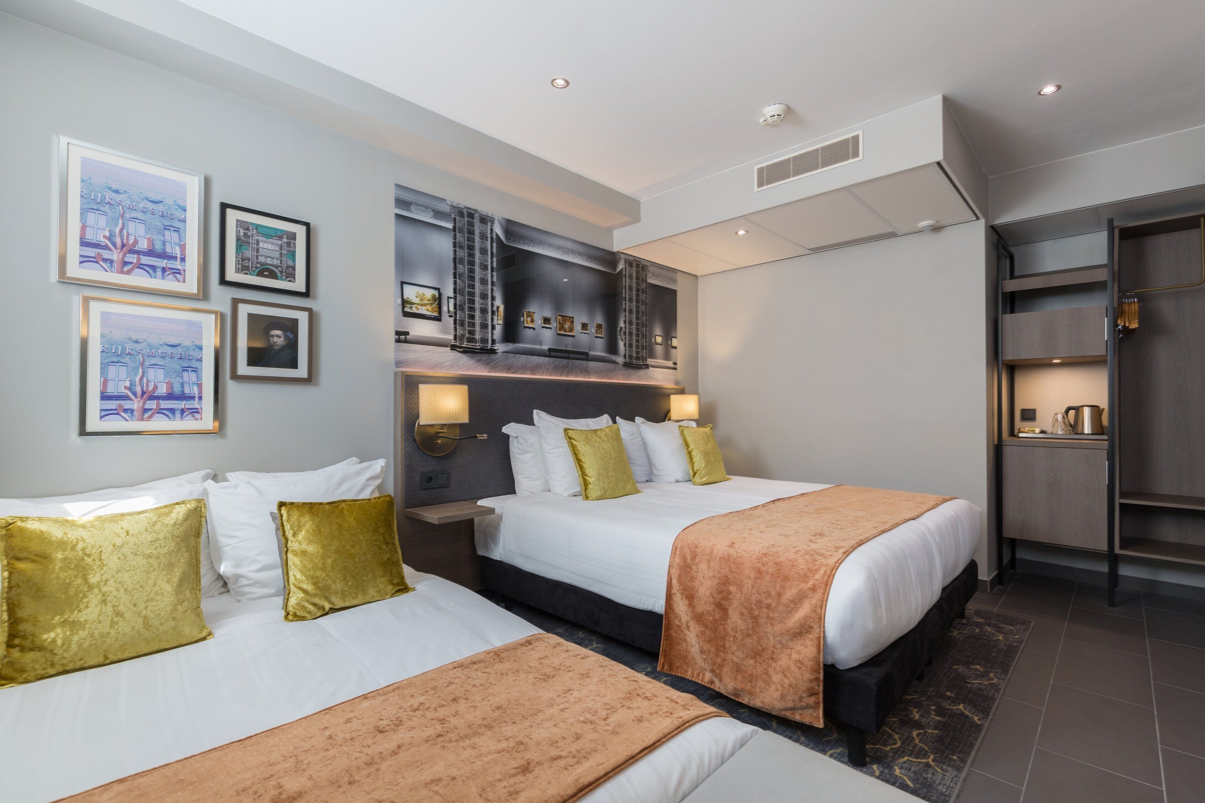 A hotel room with two beds, a large mirror, artwork on the walls, and a kitchenette.
