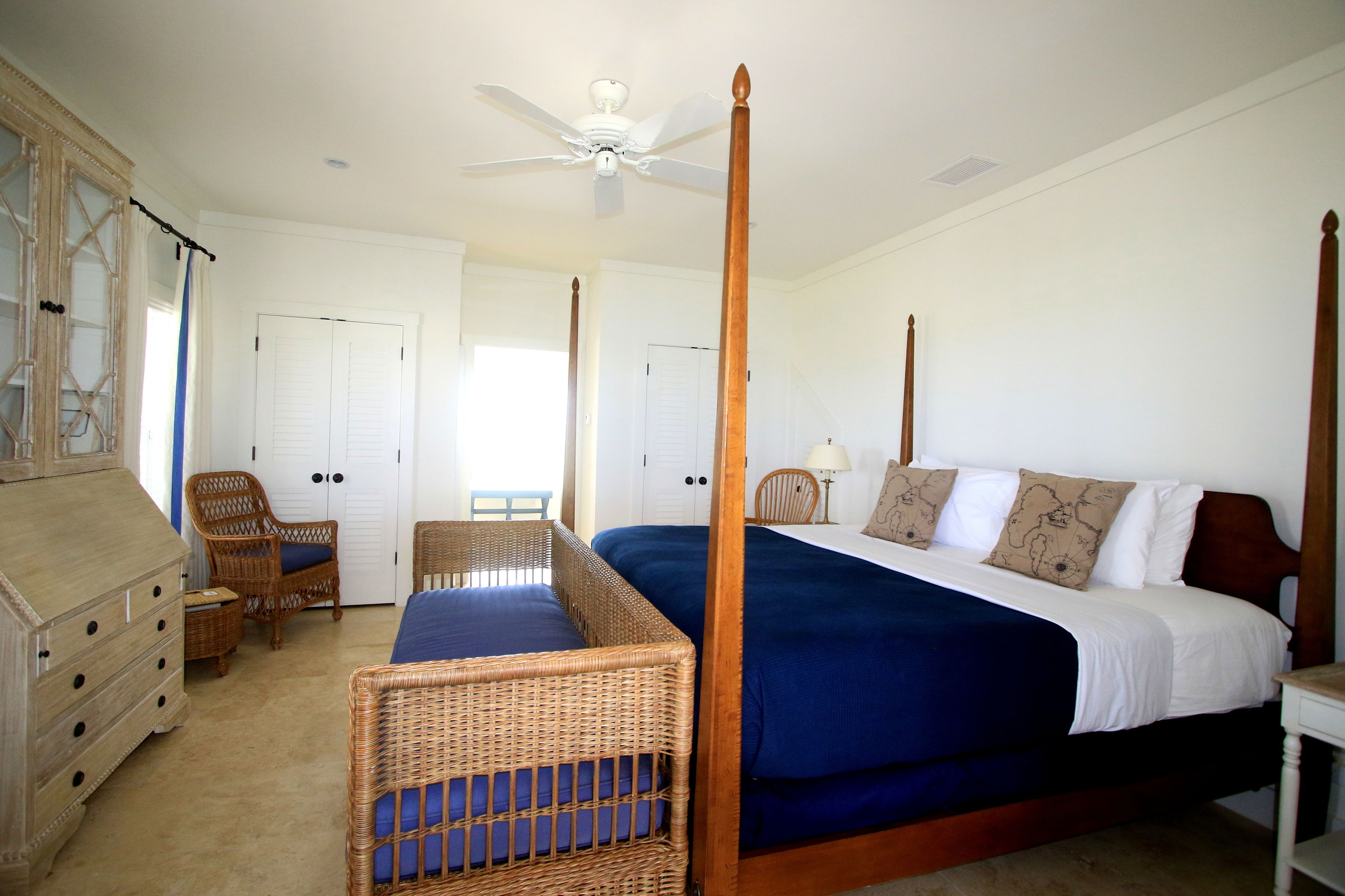 Una cama con dosel de madera, con colcha azul y dos almohadas decorativas. Se observa una silla de mimbre con cojín azul, un aparador de madera y una puerta blanca al fondo.