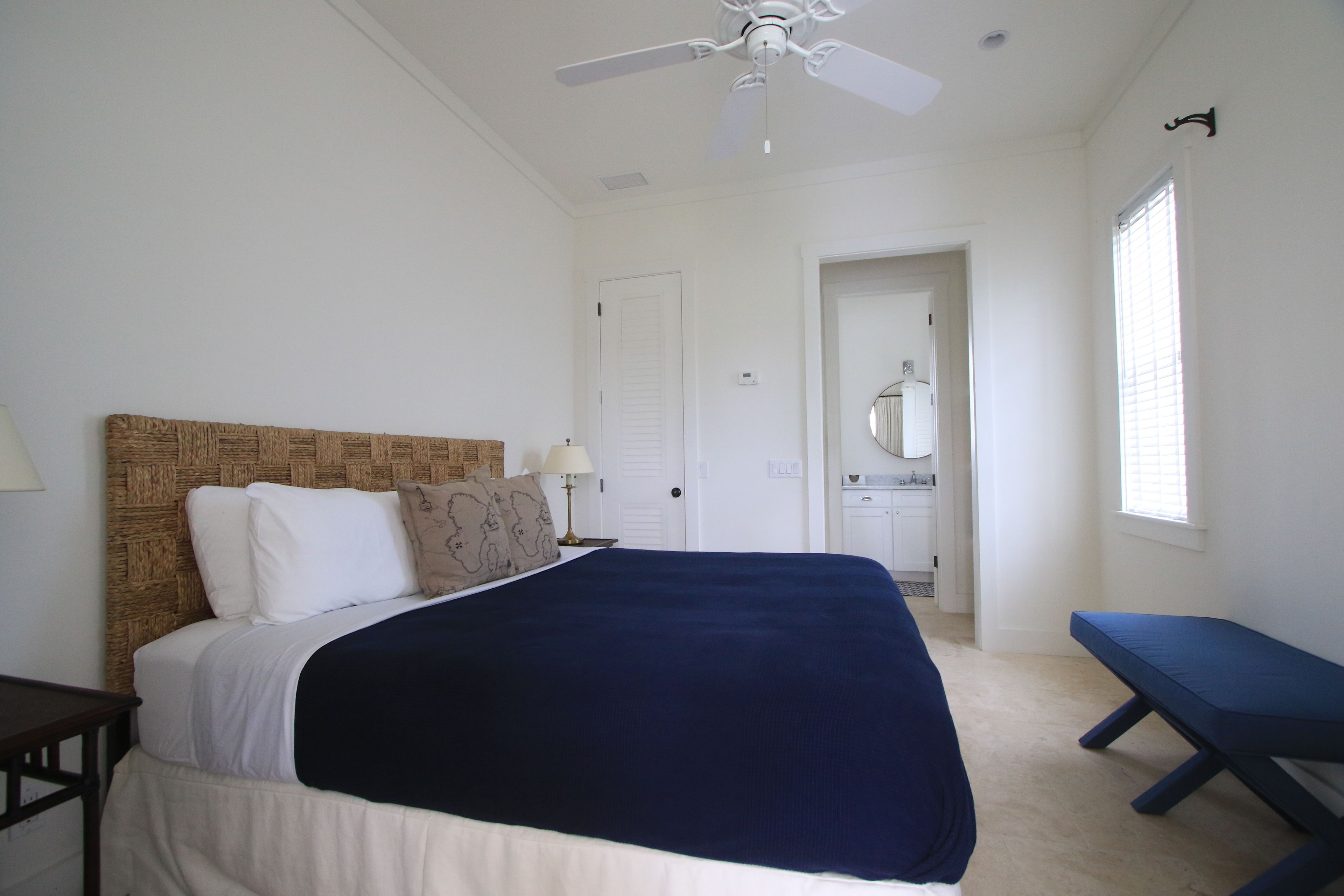 A bedroom with a bed, a bedside table, a ceiling fan, a window with blinds, and a bathroom visible through an open door.