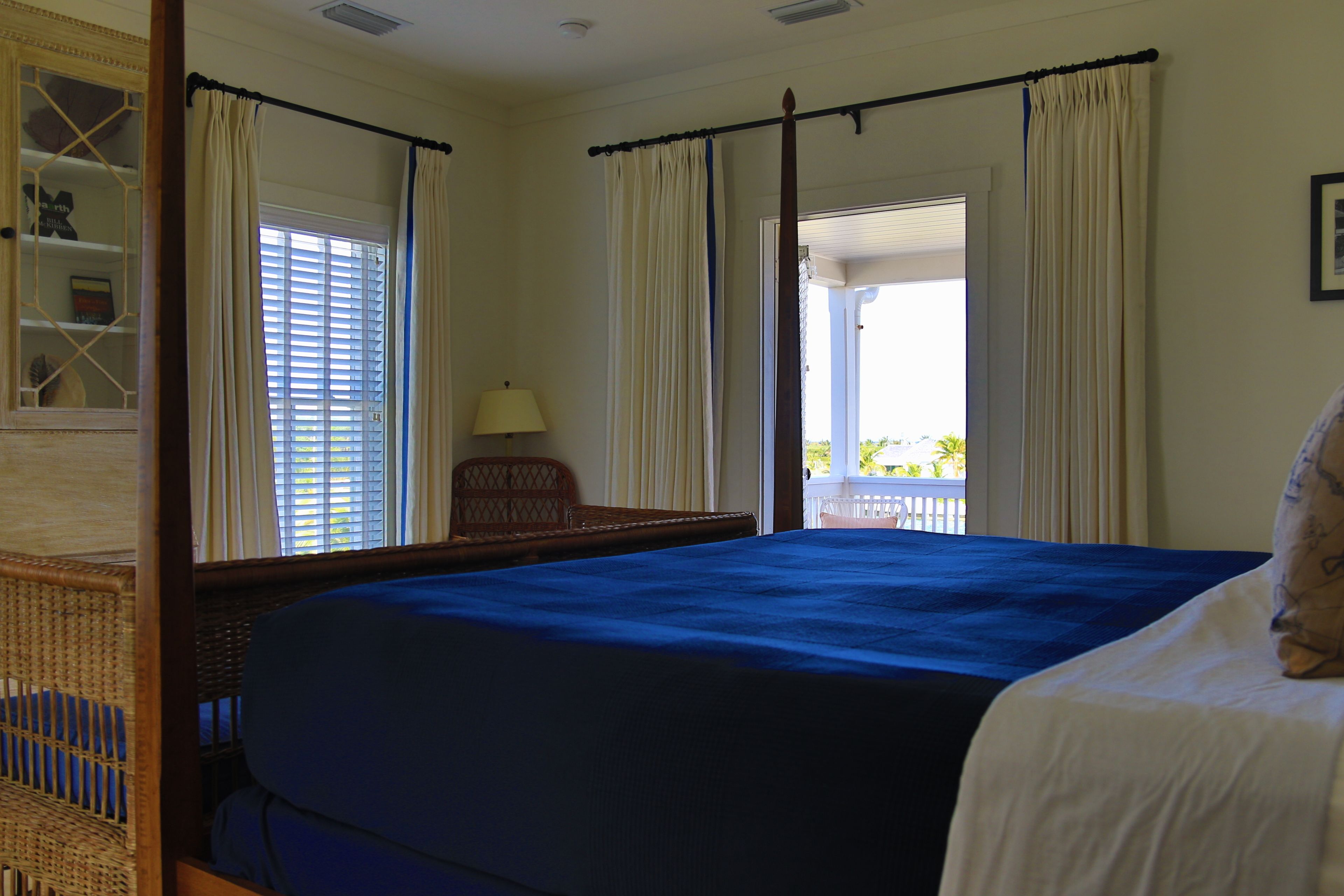 Ein Bett mit Baldachin, mit blauer Bettdecke, ein Korbstuhl, eine Lampe und ein Ausblick ins Freie durch ein Fenster mit Vorhängen.