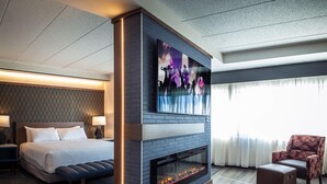 Une chambre d’hôtel moderne dotée d’un grand lit, d’un téléviseur à écran plat fixé au-dessus d’une cheminée, d’un coin salon avec un fauteuil et d’une vue sur l’extérieur par une fenêtre.