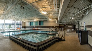 Une grande piscine intérieure avec un jacuzzi, entourée d’espaces de détente et dotée d’un haut plafond laissant apparaître les poutres de soutien.