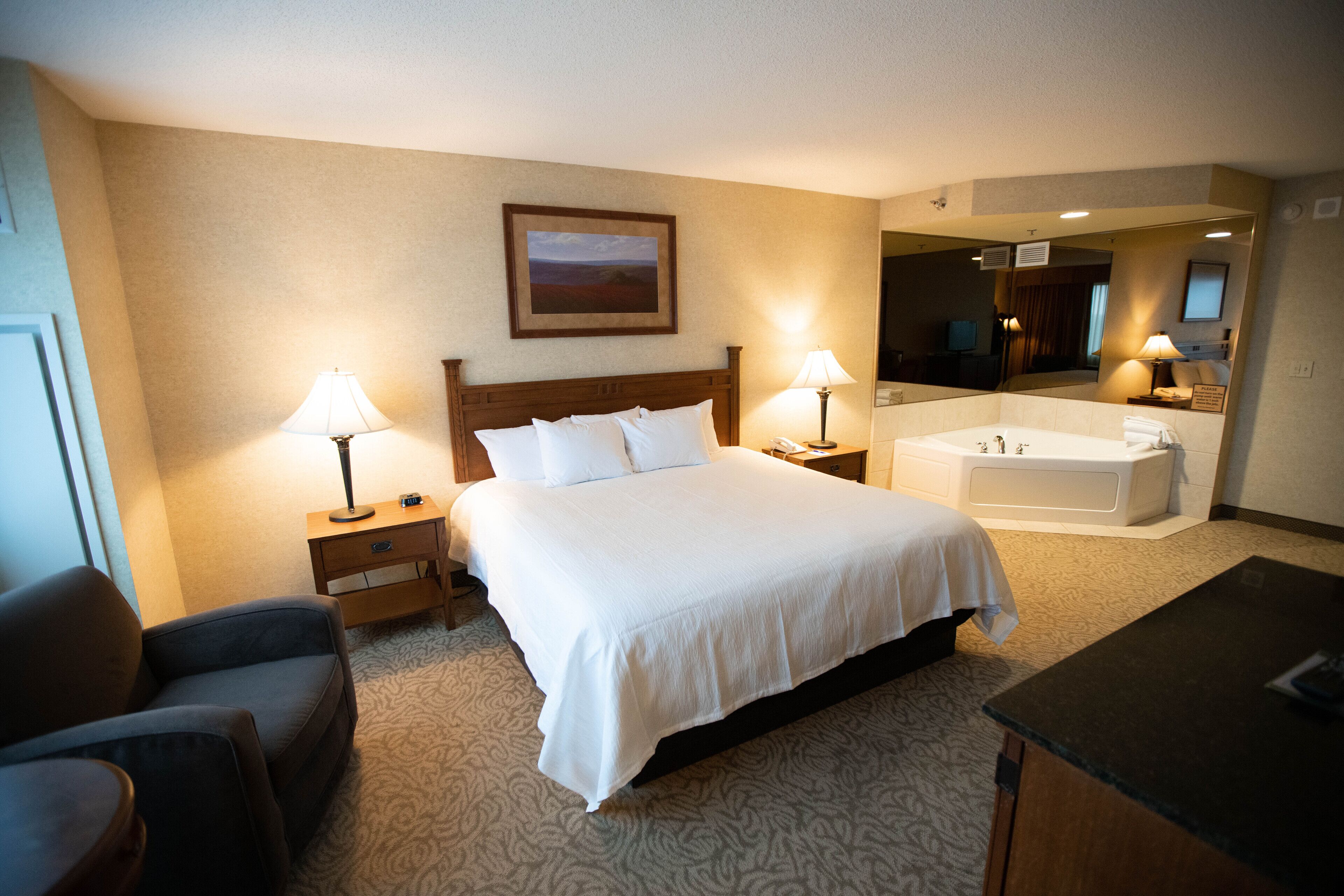 A hotel room with a large bed, two bedside tables with lamps, a sofa, a desk, a bathtub, and a mirror.