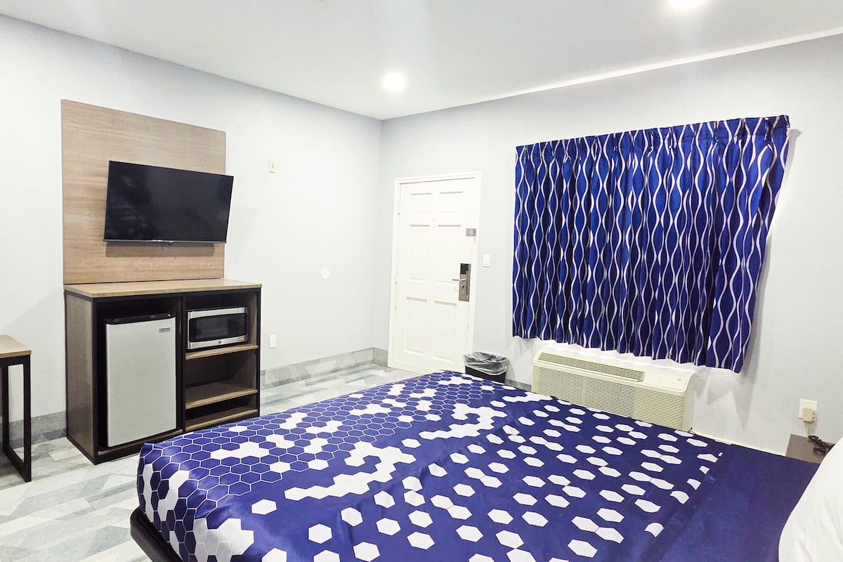 A hotel room with a bed, a desk with a chair, a TV mounted on the wall, and a window with curtains.