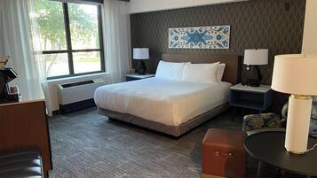 A hotel room with a large bed, two bedside tables with lamps, a small round table, a brown ottoman, and a window with curtains.
