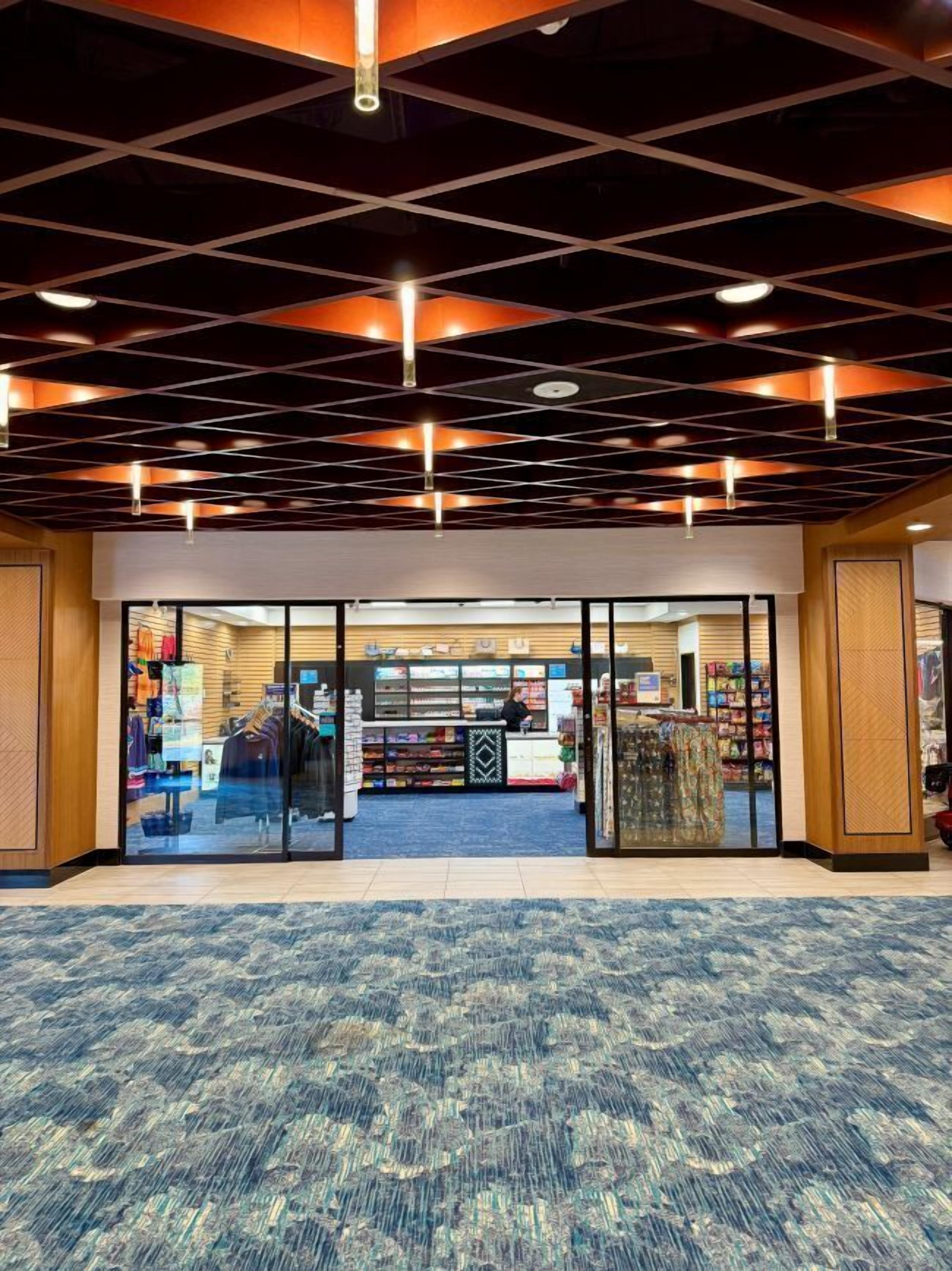 A spacious lobby with a patterned carpet, a checkered ceiling, and a view into a retail area with various products.