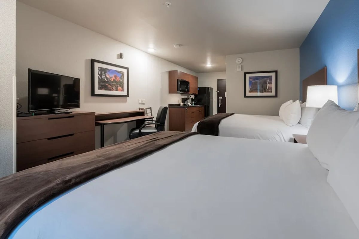 A hotel room with two beds, a desk, a television, and a kitchenette.
