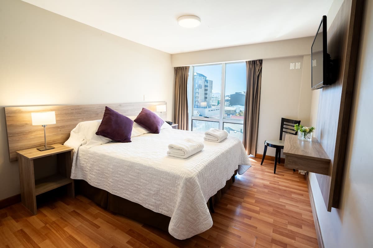Habitación de hotel con una cama grande, un escritorio, una silla, un televisor y una ventana con vistas a la ciudad.