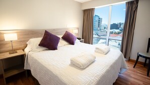 Un cuarto de hotel con una cama grande, dos almohadas, una mesita de noche con lámpara, una silla y vistas a la ciudad a través de amplios ventanales.