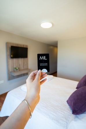 A hand holding a black card with white text in a hotel room with a bed, pillows, and a mounted TV.