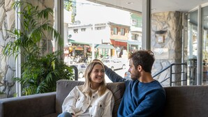 Una pareja sentada en un sofá gris en un vestíbulo moderno con una pared de piedra y ventanas amplias.