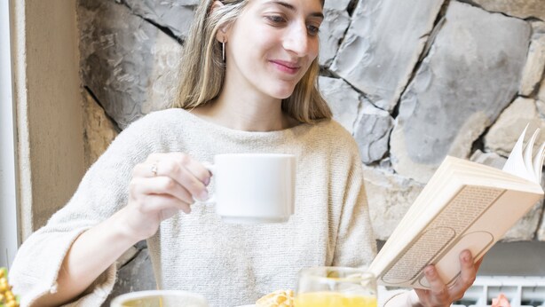 Una persona sentada a una mesa con un libro, una taza y un plato de comida que incluye tostadas y fruta.