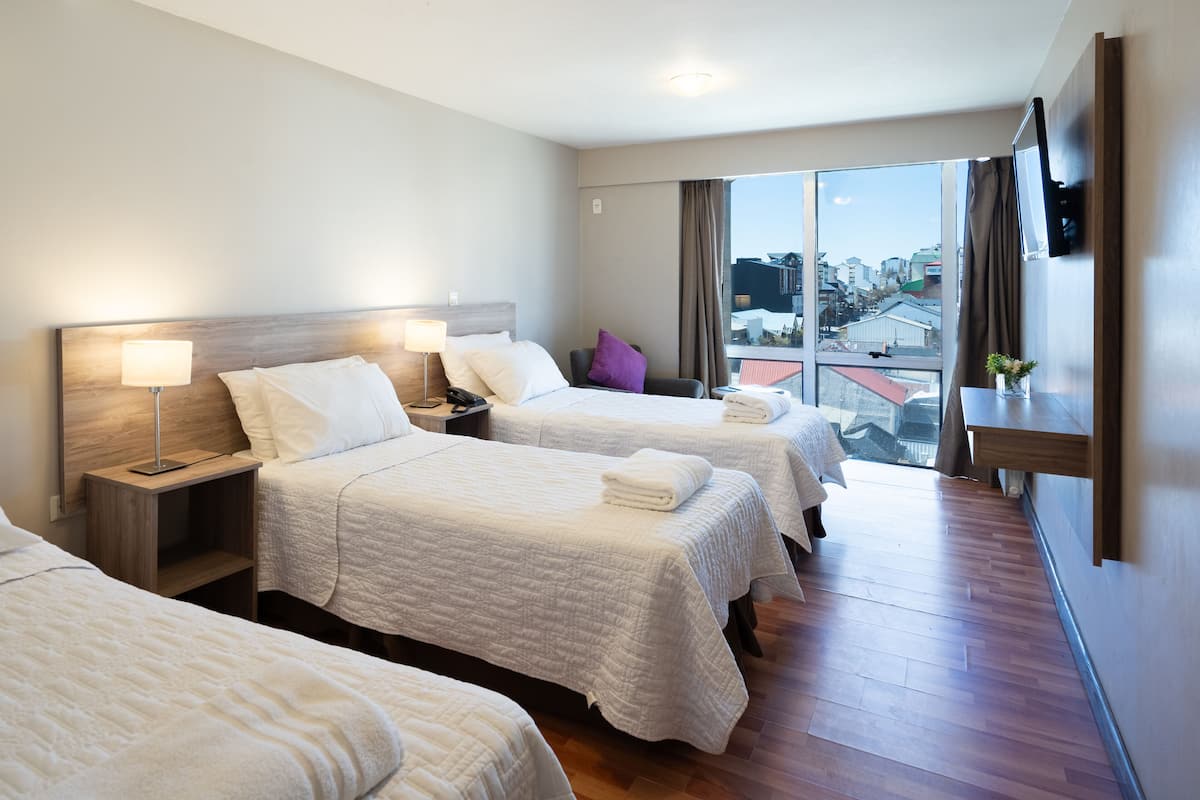 Una habitación de hotel con tres camas, un ventanal grande con vista a la ciudad, un televisor de pantalla plana montado en la pared y piso de madera.