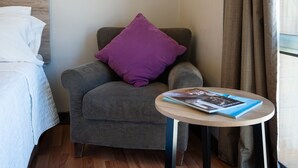 Habitación de hotel con una cama, un sillón gris con un cojín púrpura, una mesita redonda con una revista y una ventana con cortinas.